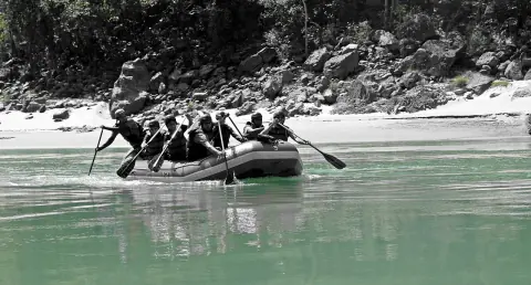 Rafters in helmets and life jackets paddling a grey raft across the green water of the Ganga