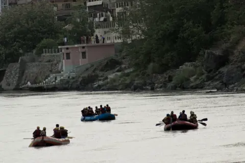 Three rafts with guests in life jackets paddling on the Ganga below the buildings and ghat steps of Rishikesh