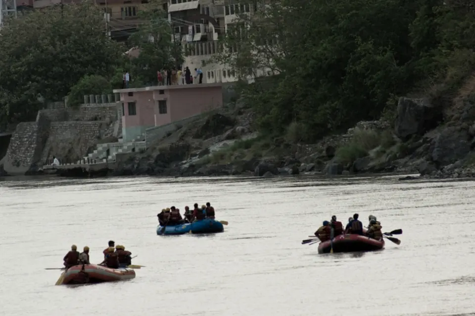 Three rafts with guests in life jackets paddling on the Ganga below the buildings and ghat steps of Rishikesh