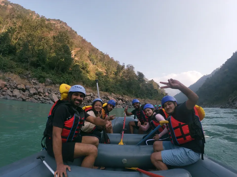 Smiling group of rafters in blue helmets and orange life jackets sitting in a raft on the Ganga with forested hills behind