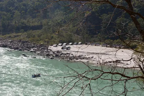 Riverside tents on a sandy beach beside the Ganga with a raft passing on the green water